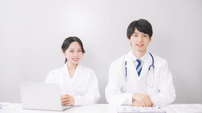 明るいオフィスのデスクで白衣を着た若い男性医師と女性医師が笑顔を見せる様子