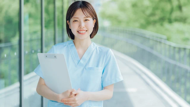 水色のスクラブを着てクリップボードを持ち、明るい渡り廊下でやさしく微笑む女性看護師